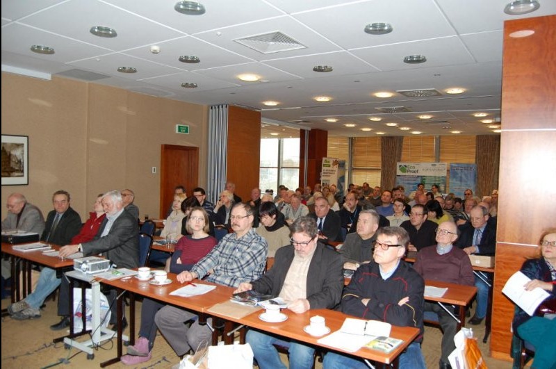 Sala konferencyjna wypełniona uczestnikami podczas prezentacji o izolacji. Widoczne materiały informacyjne i projektor na stole. Jasne oświetlenie sufitowe.