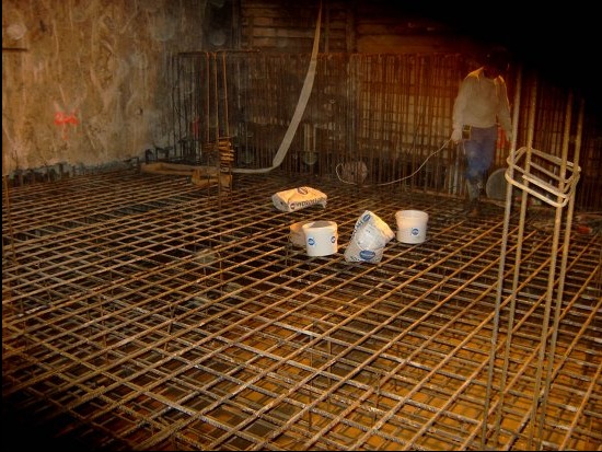 Zbrojenie fundamentu z siatki stalowej, worki z zaprawą i pracownik w tle. Przygotowanie do wylewki betonowej. Widok z góry na plac budowy.