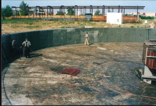 Okrągły fundament w trakcie budowy. Widoczne ściany betonowe, podłoże z śladami po pracach, pracownicy i narzędzia. W tle konstrukcja stalowa i zieleń.