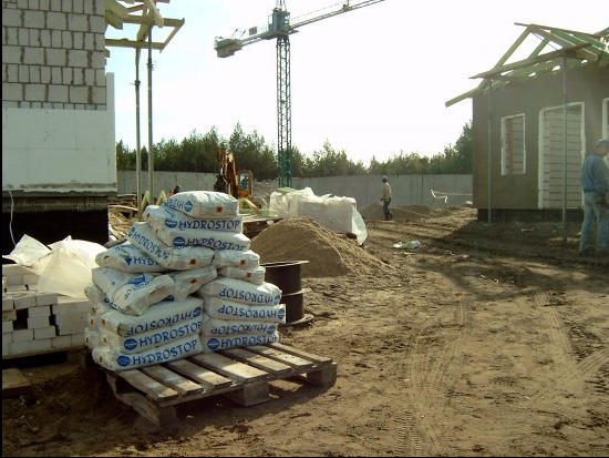 Stos worków z hydroizolacją Hydrostop na palecie, w tle budowa domu w stanie surowym otwartym, żuraw budowlany i wykop ziemny.