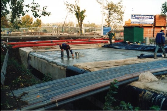 Prace na budowie: robotnik w trakcie uszczelniania fundamentów. Widoczne rury, zbiorniki i tablica informacyjna. Praca w toku, etap budowy.