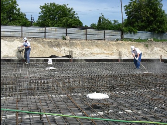 Zbrojenie fundamentów pod budowę domu jednorodzinnego. Dwóch pracowników w kaskach rozprowadza beton. Widoczne siatki zbrojeniowe i folia. Tło: ogrodzenie i drzewa.