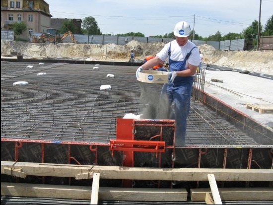 Pracownik w kasku rozsypuje materiał na zbrojenie fundamentu. Widoczne szalunki, siatka zbrojeniowa i budynek w tle. Praca na budowie w słoneczny dzień.