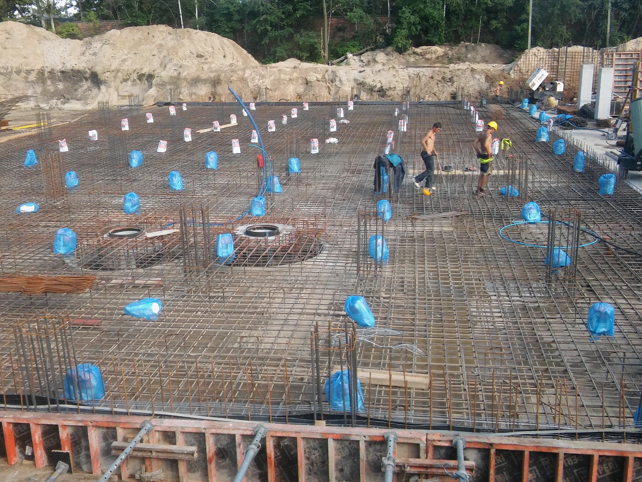 Zbrojenie fundamentów z widocznymi prętami, siatką i niebieskimi workami zabezpieczającymi, przygotowane do zalania betonem. W tle hałda piasku i pracujący robotnicy.