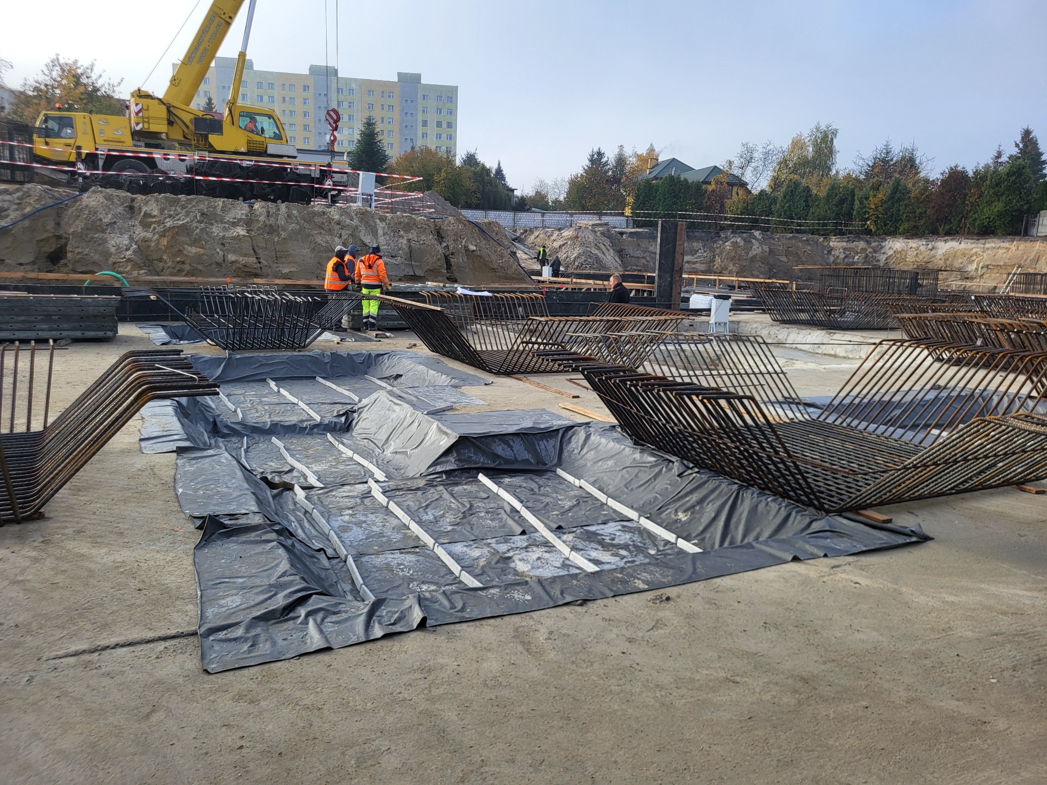 Budowa fundamentów: zbrojenie, izolacja i pracownicy w pomarańczowych kamizelkach na placu budowy z dźwigiem w tle. Widok na przygotowanie pod wylewkę.