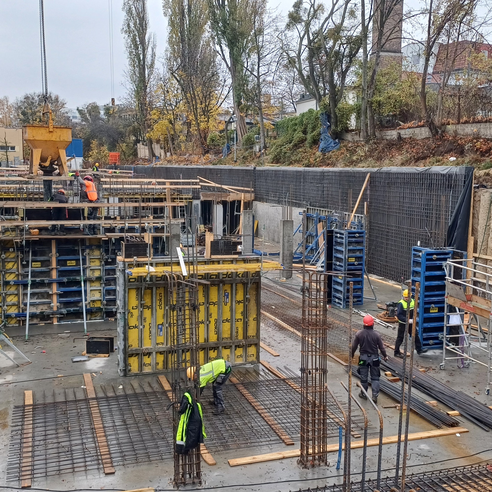 Budowa fundamentów: pracownicy w kaskach i kamizelkach układają zbrojenie, widoczne szalunki Doka, dźwig z betonem w tle, jesienna sceneria.
