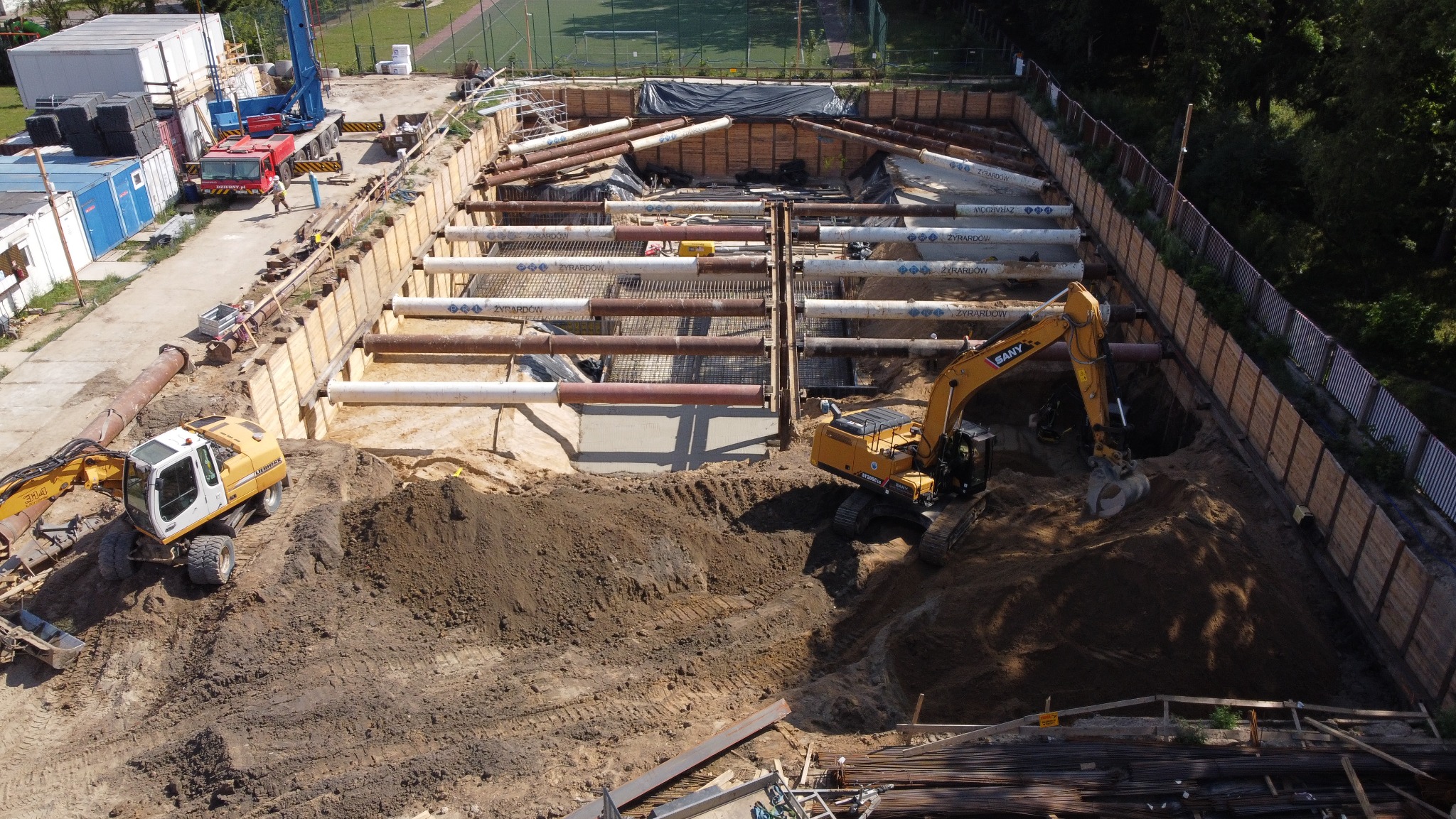 Głęboki wykop zabezpieczony szalunkiem stalowym i drewnianym, z widocznymi rurami i pracującą koparką SANY, przygotowany pod fundamenty budynku.