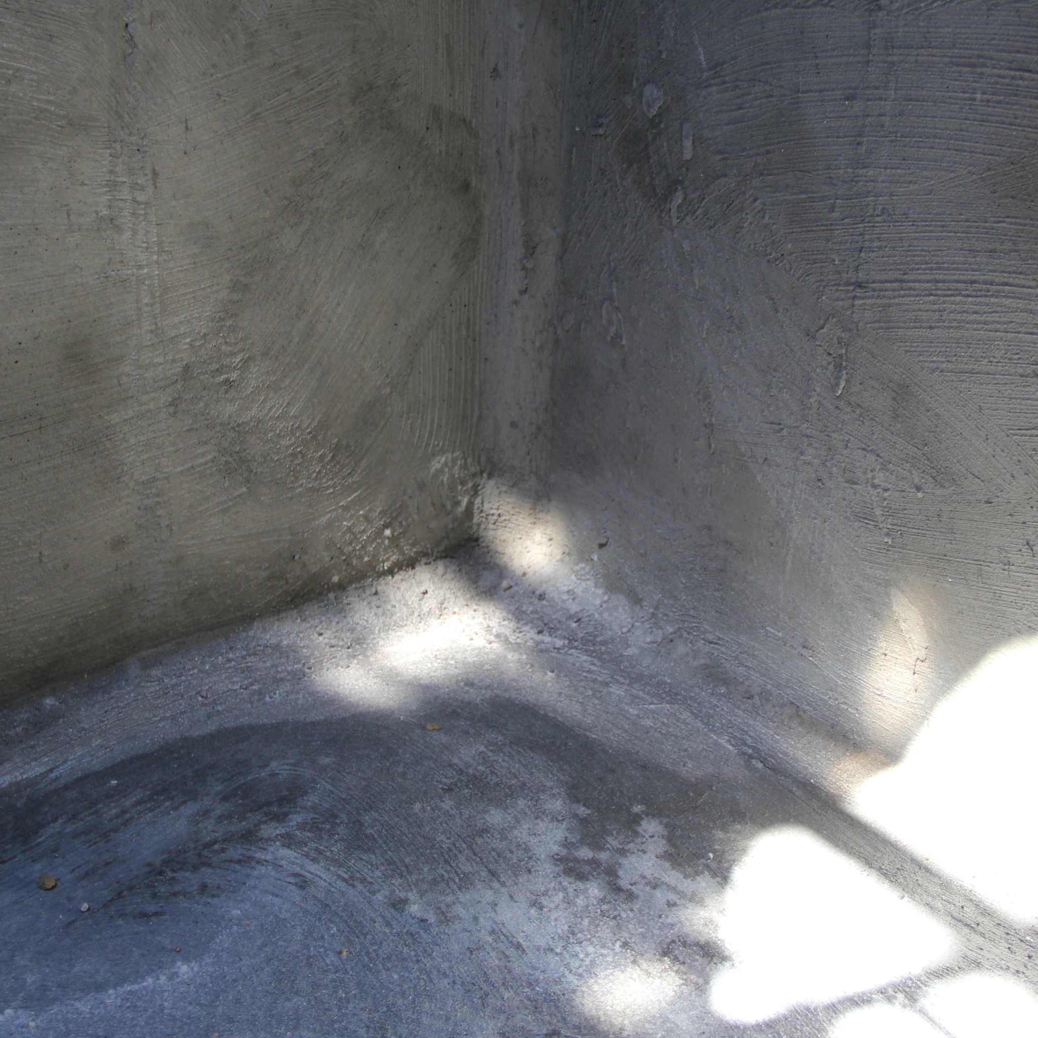 Szary, surowy narożnik fundamentu z widocznymi śladami szpachlowania i plamami wilgoci, oświetlony naturalnym światłem tworzącym jasne refleksy.