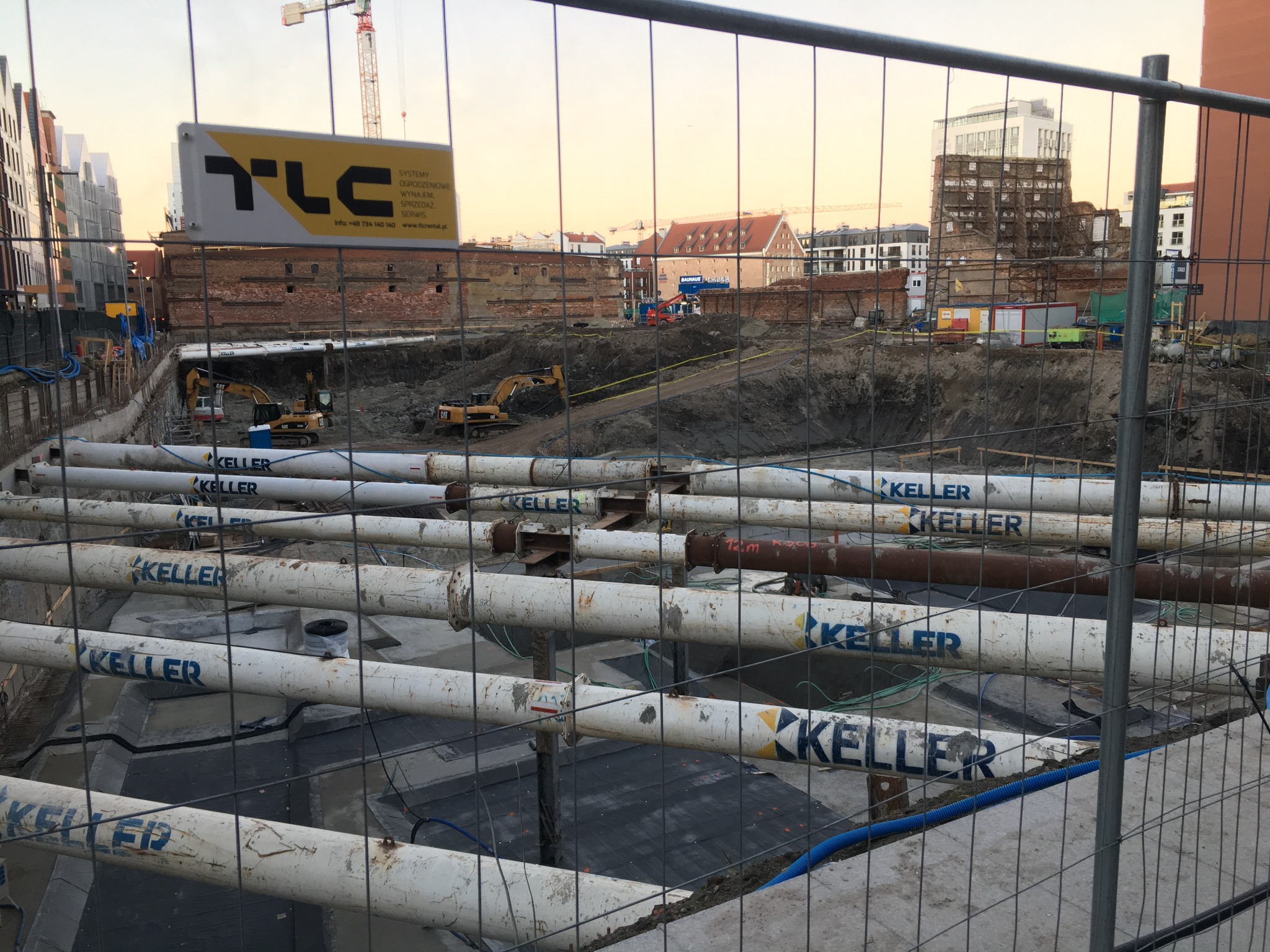 Budowa fundamentów z rurami Keller, widok przez ogrodzenie. W tle maszyny budowlane i budynki w trakcie budowy. Tablica reklamowa TLC widoczna w tle.