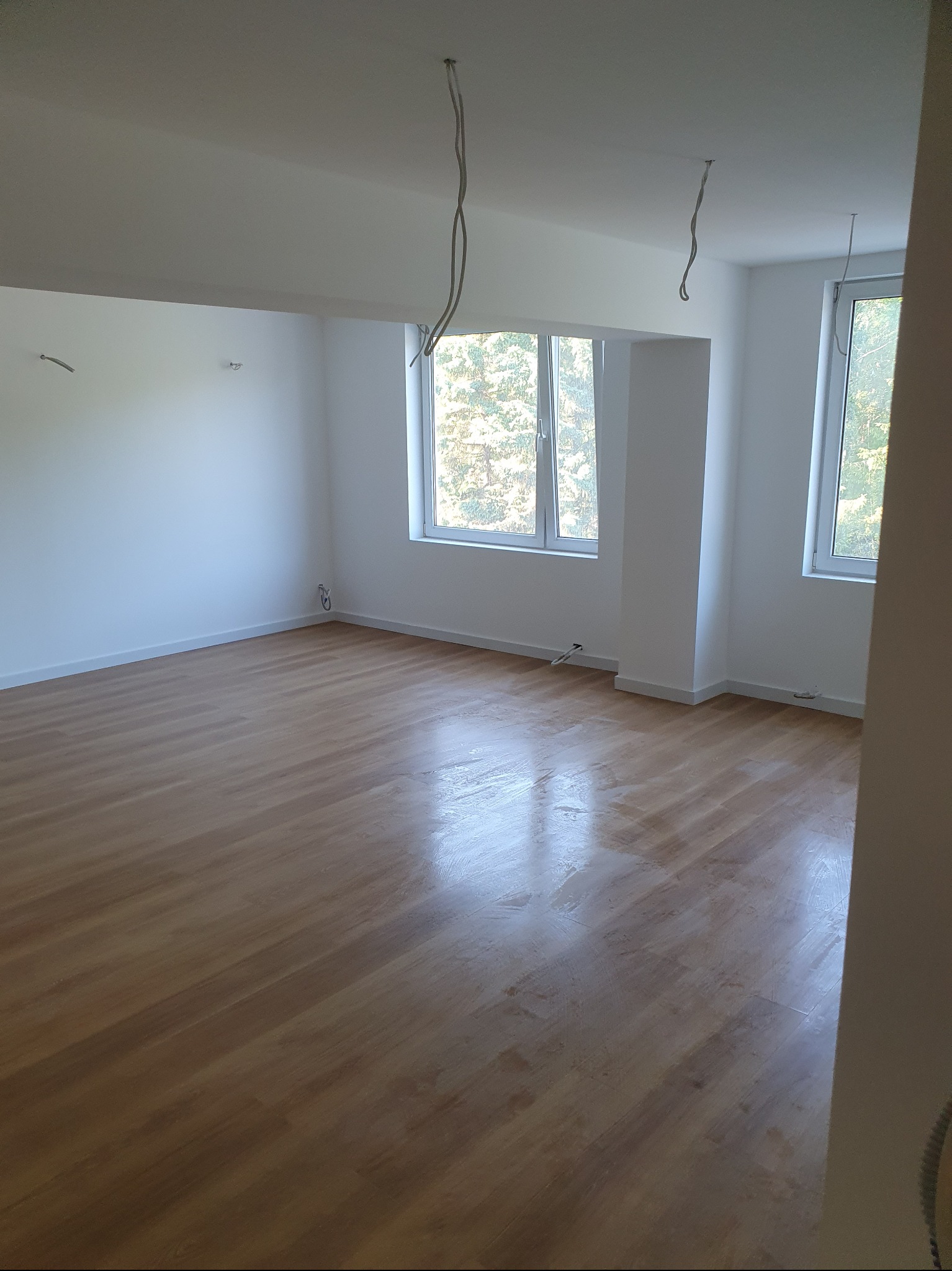 Jasne wnętrze z drewnianą podłogą i białymi ścianami. Dwa okna wpuszczają naturalne światło. Widoczne wystające kable elektryczne z sufitu.
