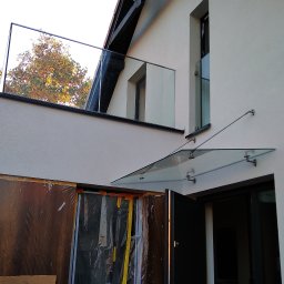 GLASS VISION MARCIN KONOPIŃSKI - Nowoczesna balustrada balkonowa ze szkła i szklany daszek nad wejściem do domu. Widoczne zabezpieczone folią okna. Jesienne drzewo w tle.