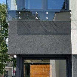 GLASS VISION MARCIN KONOPIŃSKI - Nowoczesna, szklana balustrada balkonowa z mocowaniami punktowymi, widoczna z zewnątrz budynku. Minimalistyczny design, szare elementy elewacji, refleksy w szkle.