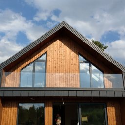 GLASS VISION MARCIN KONOPIŃSKI - Nowoczesny dom z drewnianą elewacją i balkonem zabezpieczonym szkłem, widok z dołu na tle błękitnego nieba z chmurami. Minimalistyczny design.