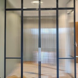 GLASS VISION MARCIN KONOPIŃSKI - Nowoczesna, wewnętrzna ścianka działowa ze szkła ryflowanego i czarnych profili, zamontowana na parkiecie. Widoczne narzędzia monterskie.