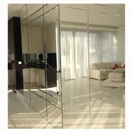 GLASS VISION MARCIN KONOPIŃSKI - Szklana ściana działowa z lustrzanych paneli w nowoczesnym wnętrzu. Widoczne odbicie kuchni i salonu z białymi meblami. Promienie słoneczne na podłodze.