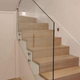 GLASS VISION MARCIN KONOPIŃSKI - Jasne, drewniane schody z przezroczystą balustradą szklaną, mocowaną na srebrnych uchwytach. Widok z dołu, z parkietem ułożonym w jodełkę i białymi szafkami pod schodami.