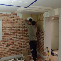 LEANDER S.C SYLWIA MATUSIEWICZ LESZEK MATUSIEWICZ - Montaż dekoracyjnej ściany z cegły w pomieszczeniu. Widoczny pracownik w trakcie układania, poziomica, wiadro z zaprawą i częściowo wykończony sufit z płyt gipsowych.