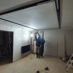 LEANDER S.C SYLWIA MATUSIEWICZ LESZEK MATUSIEWICZ - Montaż sufitu podwieszanego z płyt gipsowo-kartonowych w pomieszczeniu z instalacją elektryczną. Widoczni dwaj monterzy przy pracy, drabina i grzejnik.