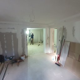 LEANDER S.C SYLWIA MATUSIEWICZ LESZEK MATUSIEWICZ - Wnętrze w trakcie prac wykończeniowych: ściany z płyt gipsowo-kartonowych, widoczne narzędzia i materiały budowlane, pracownik w oddali. Oświetlenie robocze.