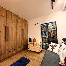 LEANDER S.C SYLWIA MATUSIEWICZ LESZEK MATUSIEWICZ - Drewniana zabudowa ściany z szafami i komodą na kółkach w pokoju w Jeleniej Górze. Na podłodze płytki imitujące drewno i niebieski dywanik.