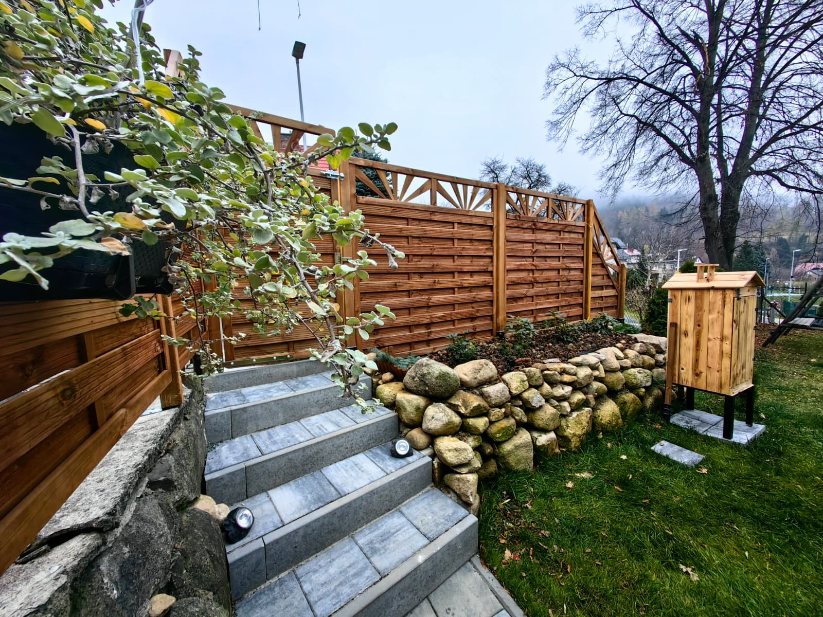 Nowoczesny ogród z drewnianym płotem, kamiennym murkiem i schodami z kostki. Widoczny drewniany domek ogrodowy i oświetlenie schodów. Całość w jesiennej scenerii.