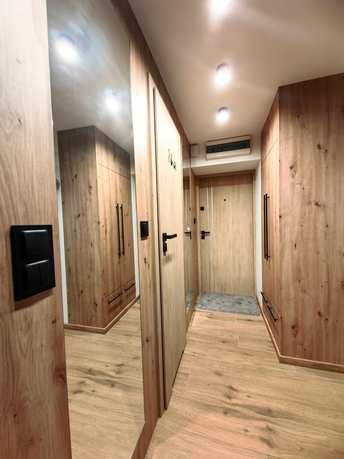 Korytarz z zabudową szaf w odcieniach drewna, lustro na ścianie odbija wnętrze. Podłoga wyłożona płytkami imitującymi drewno. Minimalistyczne oświetlenie sufitowe.