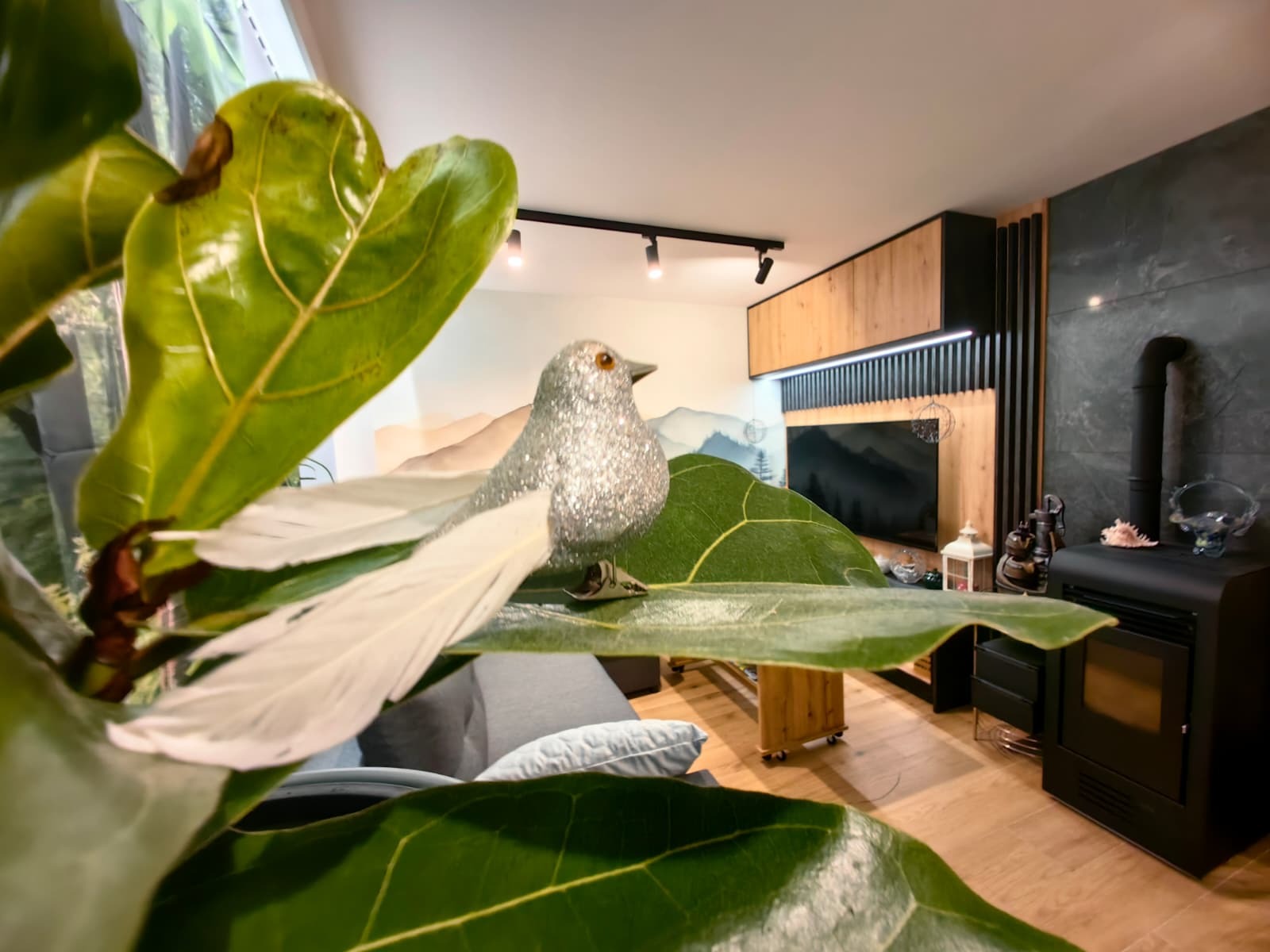 Nowoczesny salon z akcentem natury: duży liść fikusa i srebrny ptaszek na pierwszym planie, w tle ściana z telewizorem i kominkiem, drewniane elementy wykończenia.