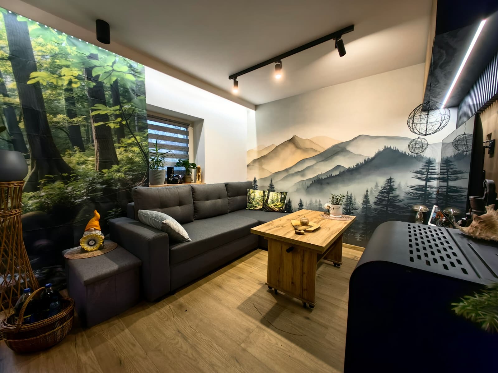 Przytulny salon z szarą sofą, drewnianym stolikiem i fototapetą z motywem górskim. Oświetlenie punktowe i dekoracje roślinne tworzą relaksującą atmosferę.