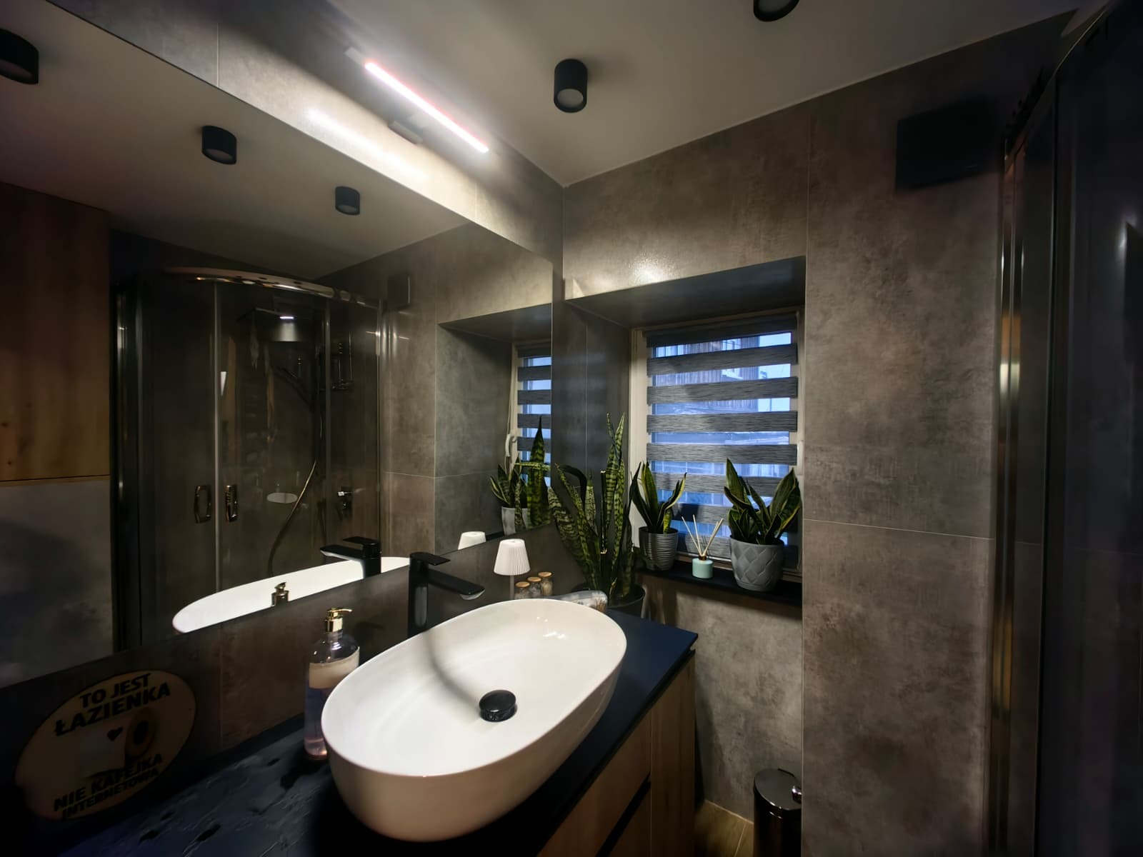 Stylowa łazienka z szarą glazurą, owalną umywalką nablatową i kabiną prysznicową. Okno z roletami dzień-noc i roślinami doniczkowymi dodaje naturalnego akcentu.