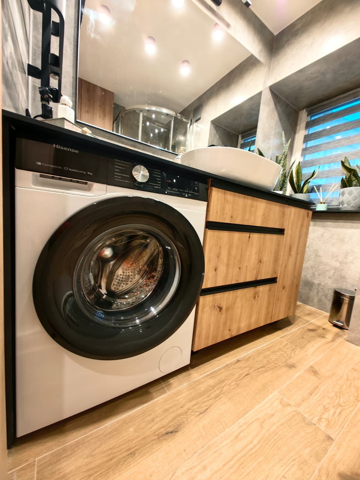 Nowoczesna łazienka z pralką Hisense w zabudowie z drewnianymi frontami, umywalką nablatową i szarą ścianą. Widoczne rośliny w oknie i drewniana podłoga.