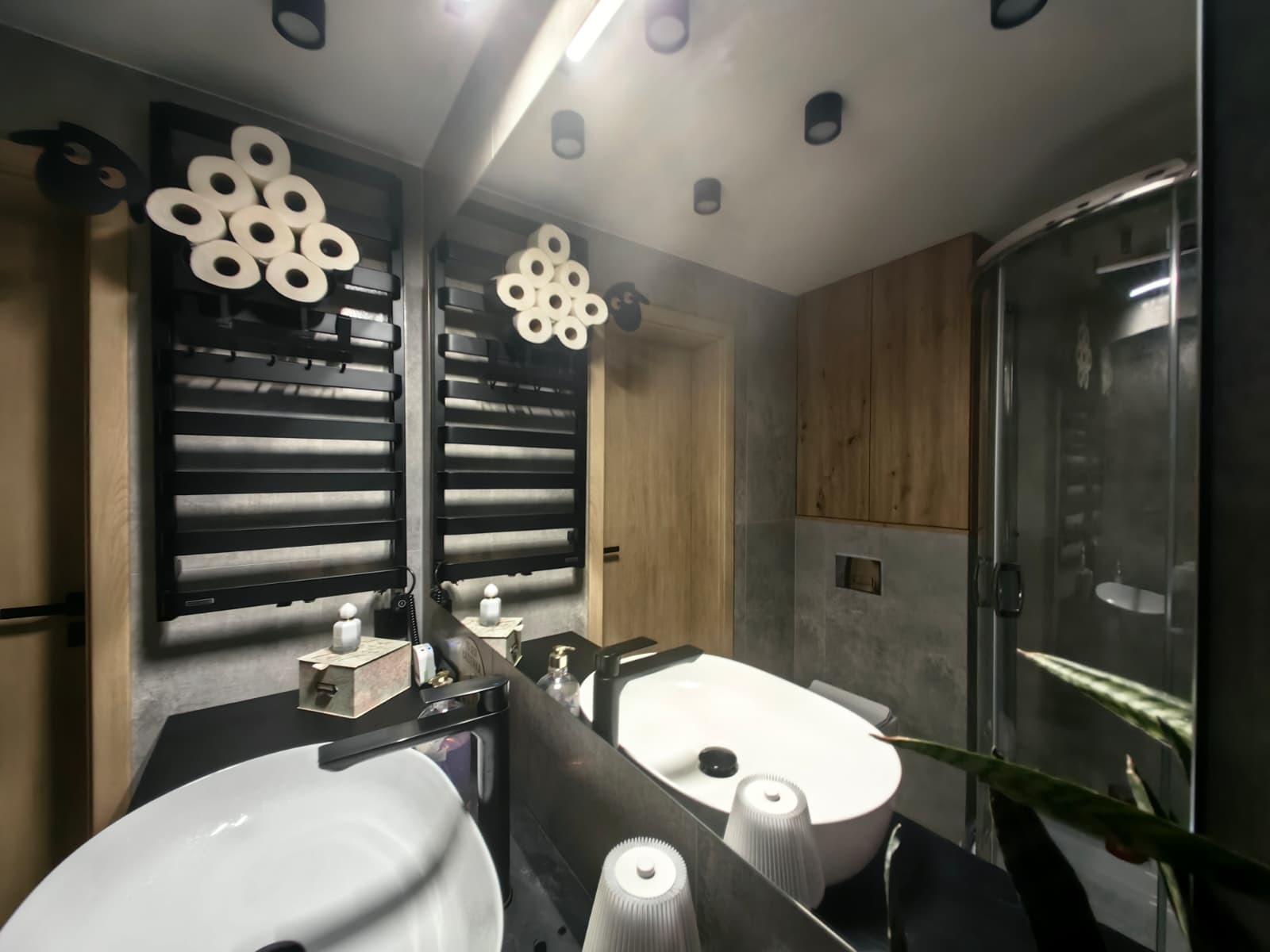 Nowoczesna łazienka z owalną umywalką, czarnym grzejnikiem drabinkowym i dekoracją z papieru toaletowego. Widoczne szklana kabina prysznicowa i drewniane szafki.