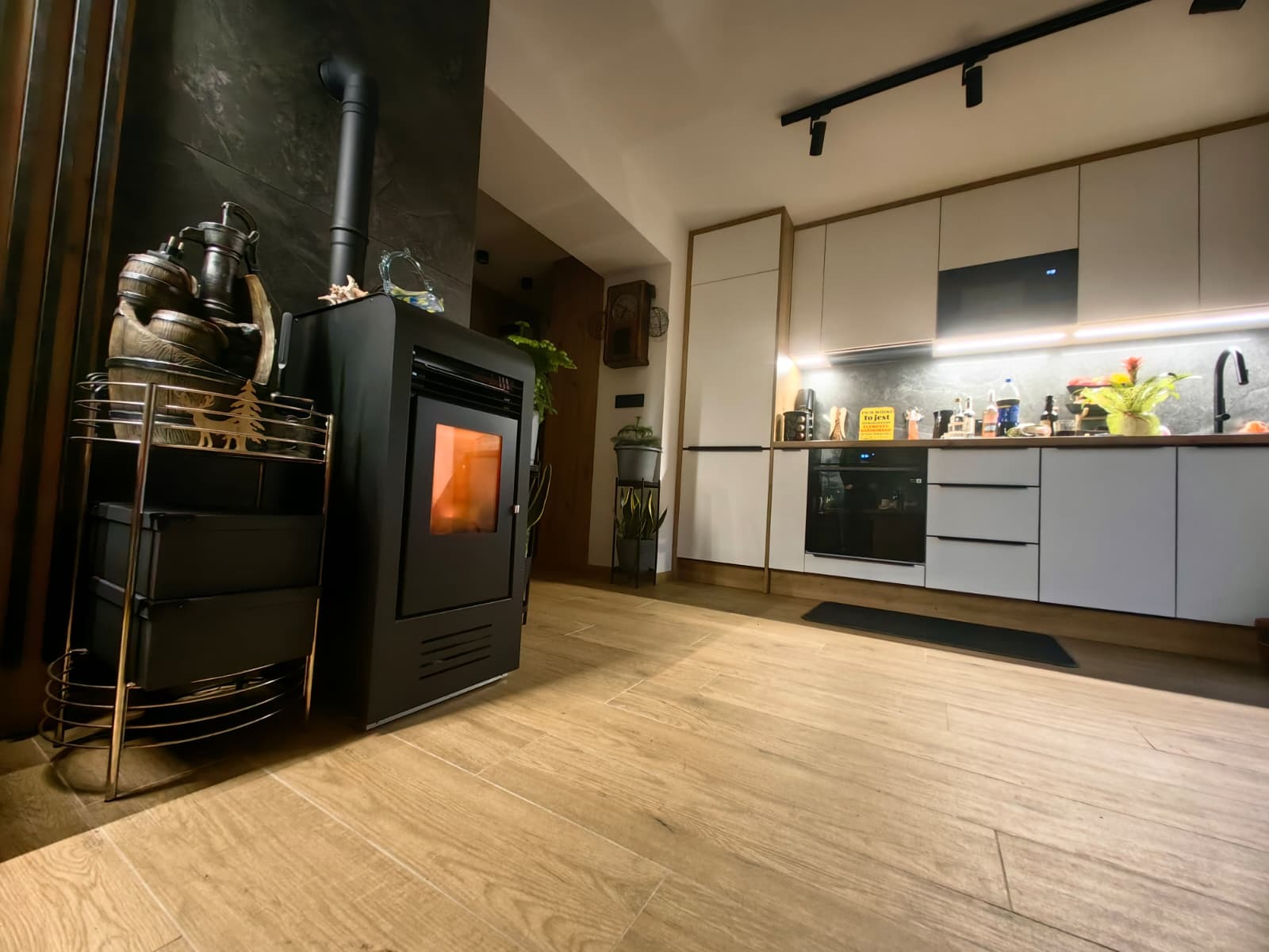 Nowoczesna kuchnia z drewnianą podłogą i czarnym piecem wolnostojącym. Białe szafki kuchenne z oświetleniem LED. Minimalistyczny design z elementami dekoracyjnymi.