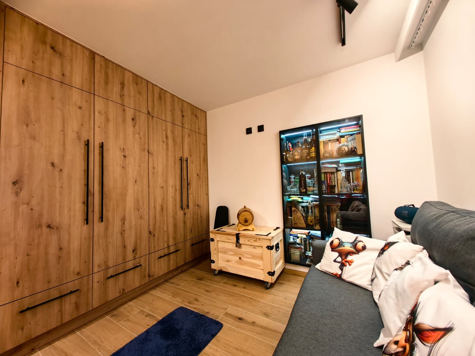 Drewniana zabudowa ściany z szafami i komodą na kółkach w pokoju w Jeleniej Górze. Na podłodze płytki imitujące drewno i niebieski dywanik.