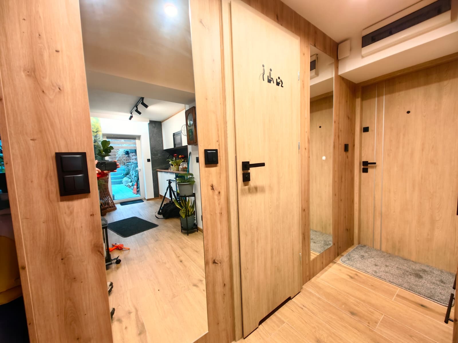 Jasne drewno i czarne akcenty w wykończeniu wnętrza: drzwi z piktogramami, podłoga z płytek imitujących drewno, widok na kuchnię i ogród. Nowoczesny design.