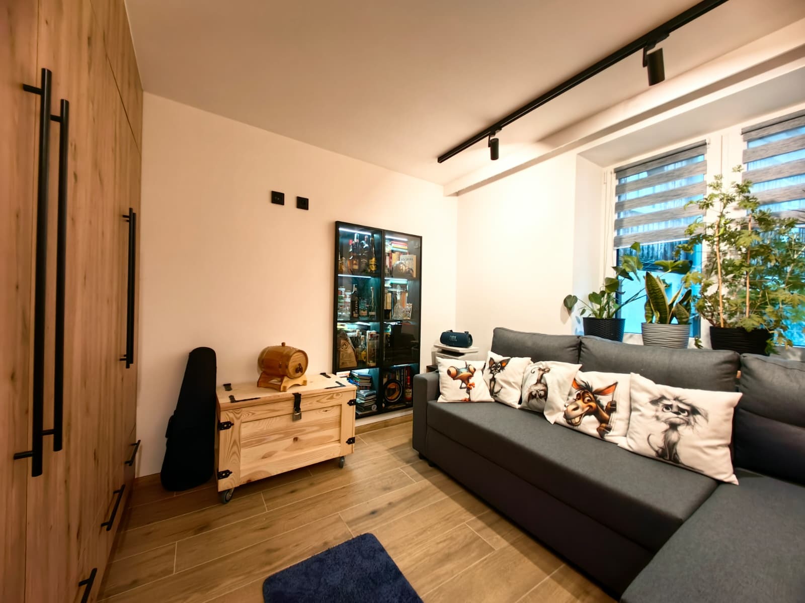 Jasne drewno szafy i podłogi w salonie z szarą sofą z poduszkami w zwierzęce wzory, skrzynią i regałem z alkoholem. Nowoczesne oświetlenie sufitowe.