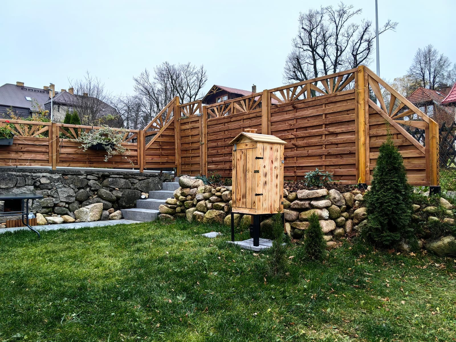 Ogród z drewnianym płotem z ozdobnymi detalami, kamiennym murkiem i schodami. Na trawniku stoi drewniana wędzarnia. Widok na domy w tle.
