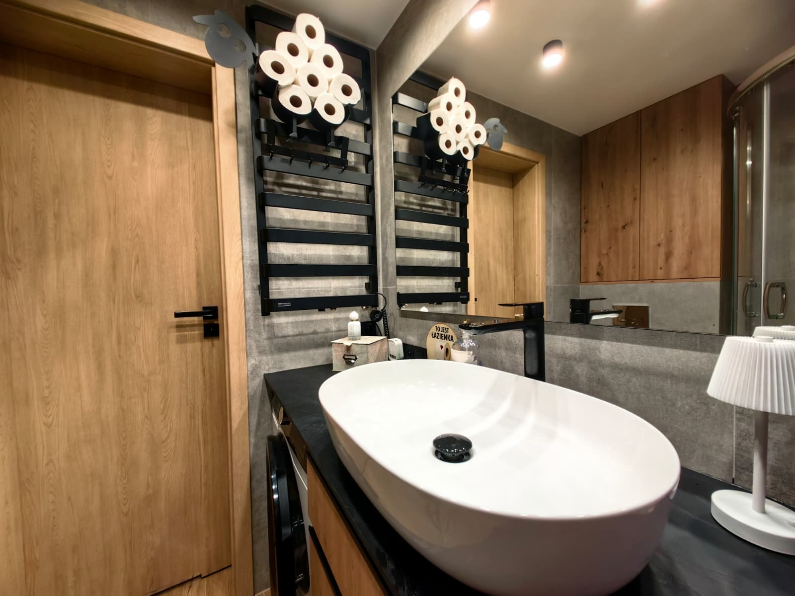 Nowoczesna łazienka z owalną umywalką nablatową, czarną armaturą i grzejnikiem drabinkowym z nietypowym uchwytem na papier toaletowy. Drewniane szafki i szara ściana.
