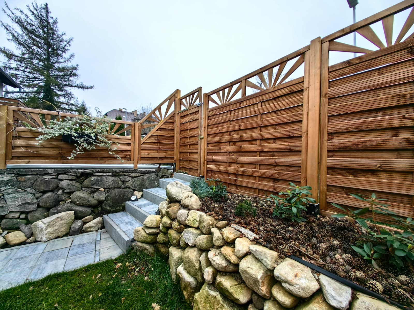 Drewniany płot z bramką, z dekoracyjnym wzorem w górnej części, otacza kamienny murek i schody. Widoczne rośliny i trawnik. Całość w naturalnej kolorystyce.