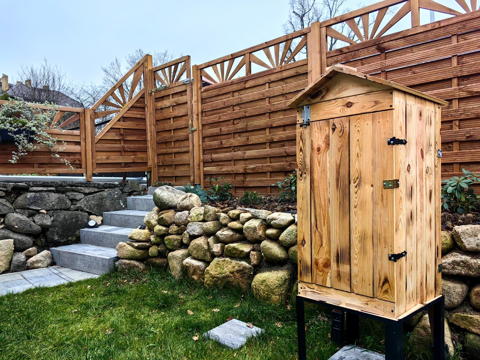 Drewniana wędzarnia ogrodowa na tle ażurowego ogrodzenia z drewna, kamiennego murku i schodów. Całość wkomponowana w zielony trawnik.