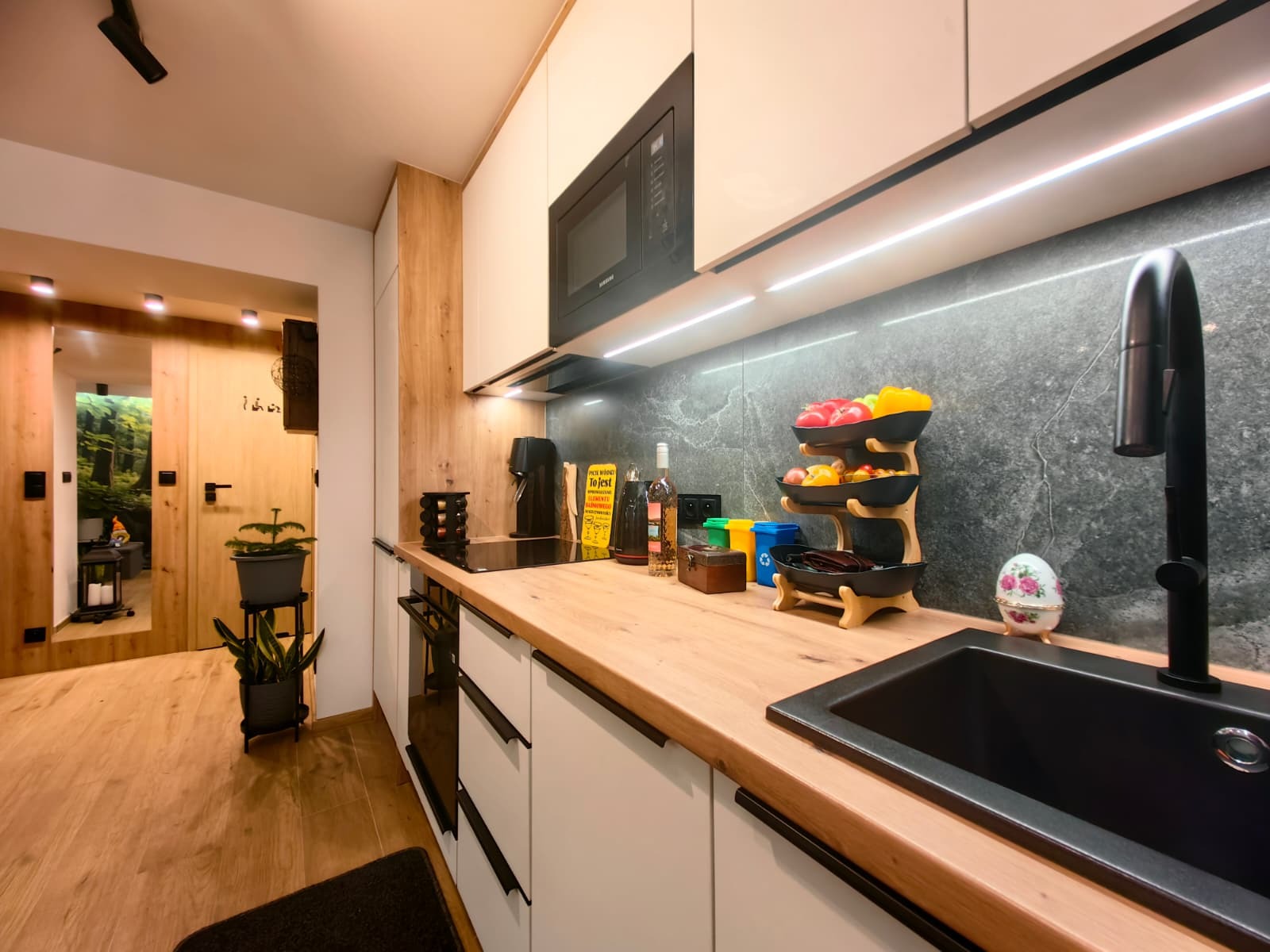 Nowoczesna kuchnia z drewnianym blatem, białymi szafkami i czarnymi akcentami. Na blacie dekoracje, owoce i czarna armatura. Widoczne oświetlenie LED i marmurowa ściana.