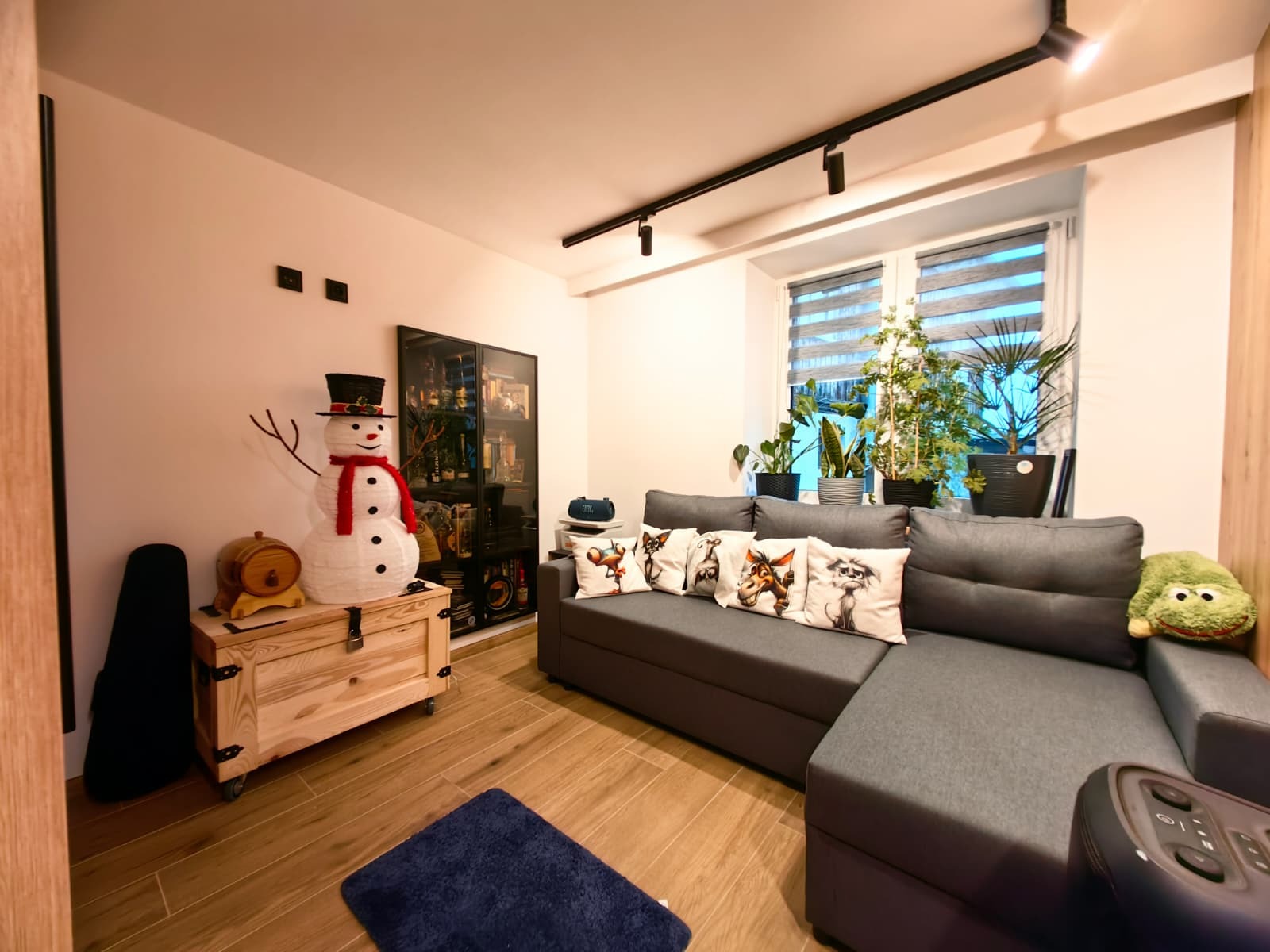 Jasne wnętrze salonu z szarą sofą, drewnianą podłogą i dekoracjami: bałwan, rośliny w oknach, szafka z oświetleniem punktowym. Przytulna aranżacja.