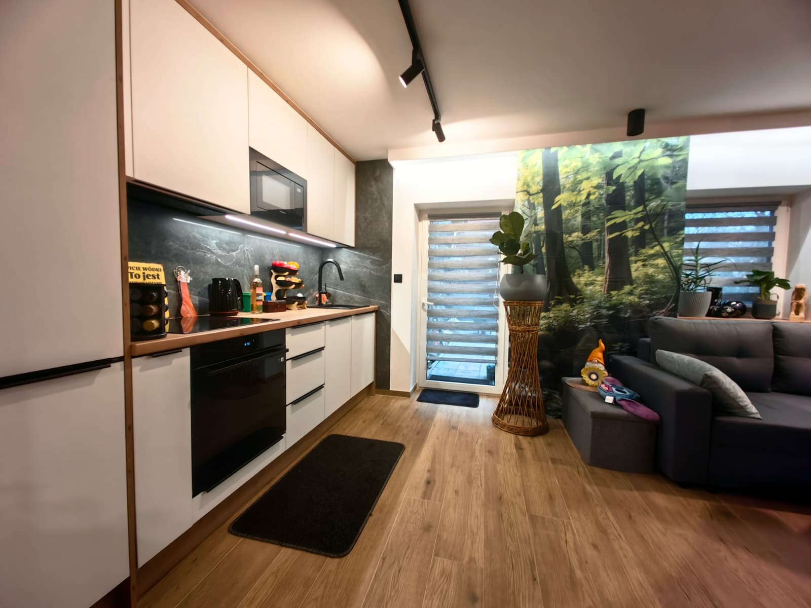 Nowoczesna kuchnia z białymi szafkami, czarnym blatem i sprzętem AGD. Na ścianie fototapeta z motywem leśnym. Drewniana podłoga i oświetlenie punktowe.