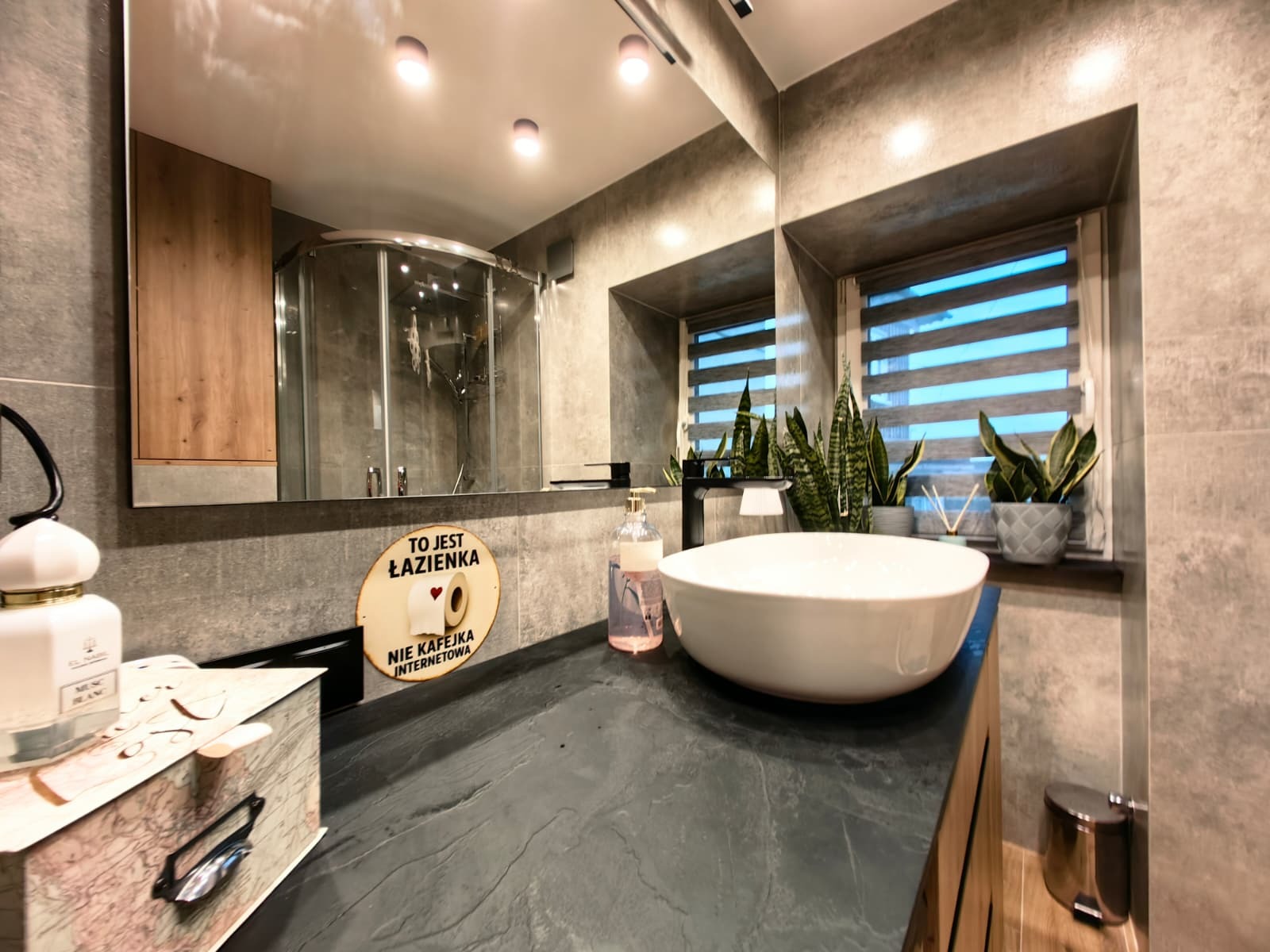 Nowoczesna łazienka z szarą glazurą, kabiną prysznicową w lustrze i umywalką nablatową. Uwagę przykuwa dekoracyjny napis 'To jest łazienka, nie kafejka internetowa'.