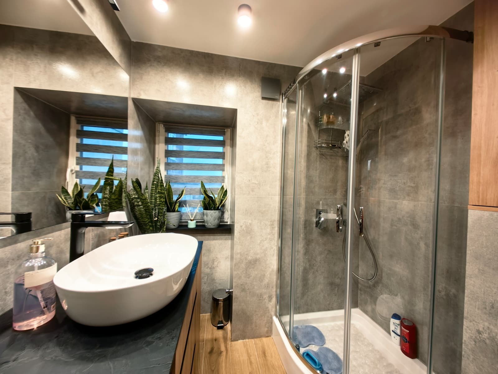 Nowoczesna łazienka z szarą glazurą, owalną umywalką na blacie z czarnego kamienia i kabiną prysznicową z zaokrąglonymi drzwiami. Na parapecie rośliny w doniczkach.