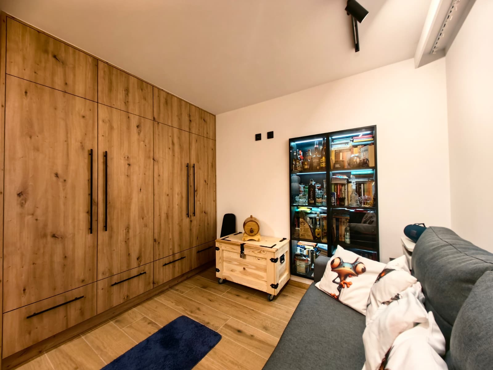 Drewniana szafa wnękowa z czarnymi uchwytami, podłoga z paneli, przeszklona witryna z podświetleniem i szara sofa w pokoju. Widok na wykończone wnętrze.