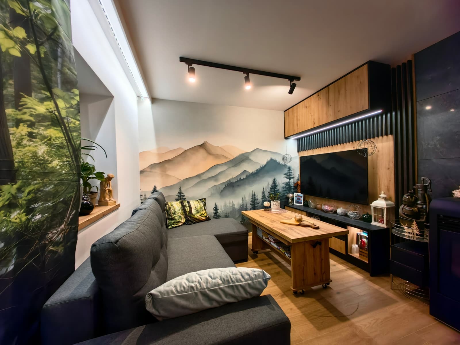 Stylowy salon z szarą sofą narożną, drewnianym stolikiem na kółkach i ścianą z motywem górskim. Nowoczesne oświetlenie i zabudowa TV z drewnianymi elementami.
