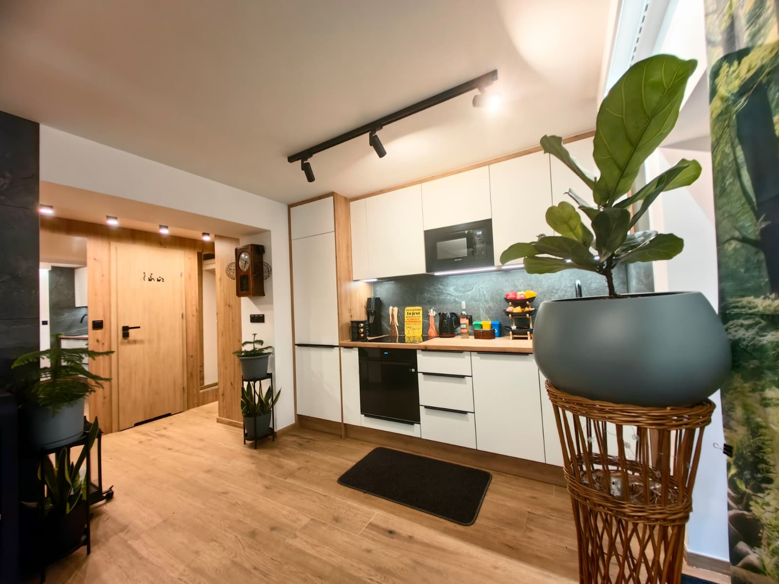 Jasna kuchnia z drewnianymi akcentami, oświetleniem punktowym i roślinami doniczkowymi. Widoczny fragment salonu z drewnianymi drzwiami i zegarem.