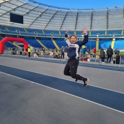 Studio treningowe Angelika Noga Power Team - Uśmiechnięta kobieta w sportowym stroju skacze na bieżni stadionu, unosząc ręce w geście radości. W tle widoczne trybuny i brama startowa.