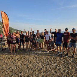 Studio treningowe Angelika Noga Power Team - Grupa biegaczy na plaży po biegu, z pomarańczową flagą 'Dalmacija Ultra Trail' w tle. Uczestnicy w strojach sportowych, niektórzy z numerami startowymi.
