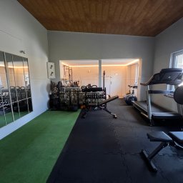 Angelika Noga Power Team - Domowa siłownia z bieżnią, ławeczką, lustrem i różnorodnym sprzętem do ćwiczeń. Podłoga wyłożona matą, sufit drewniany.